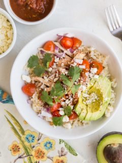 Chicken burrito bowl with avocado, salsa, rice, cheese, and shredded chicken, with a flower napkin and avocado to the side and bowls of cheese and salsa.