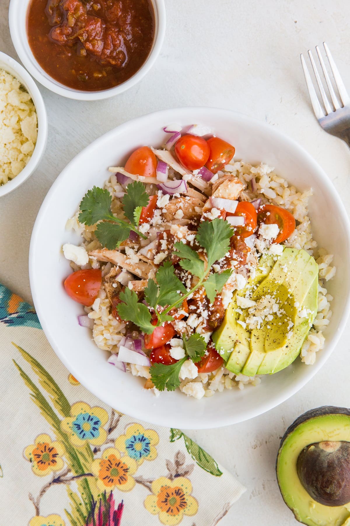 Chicken burrito bowl with avocado, salsa, rice, cheese, and shredded chicken, with a flower napkin and avocado to the side and bowls of cheese and salsa.