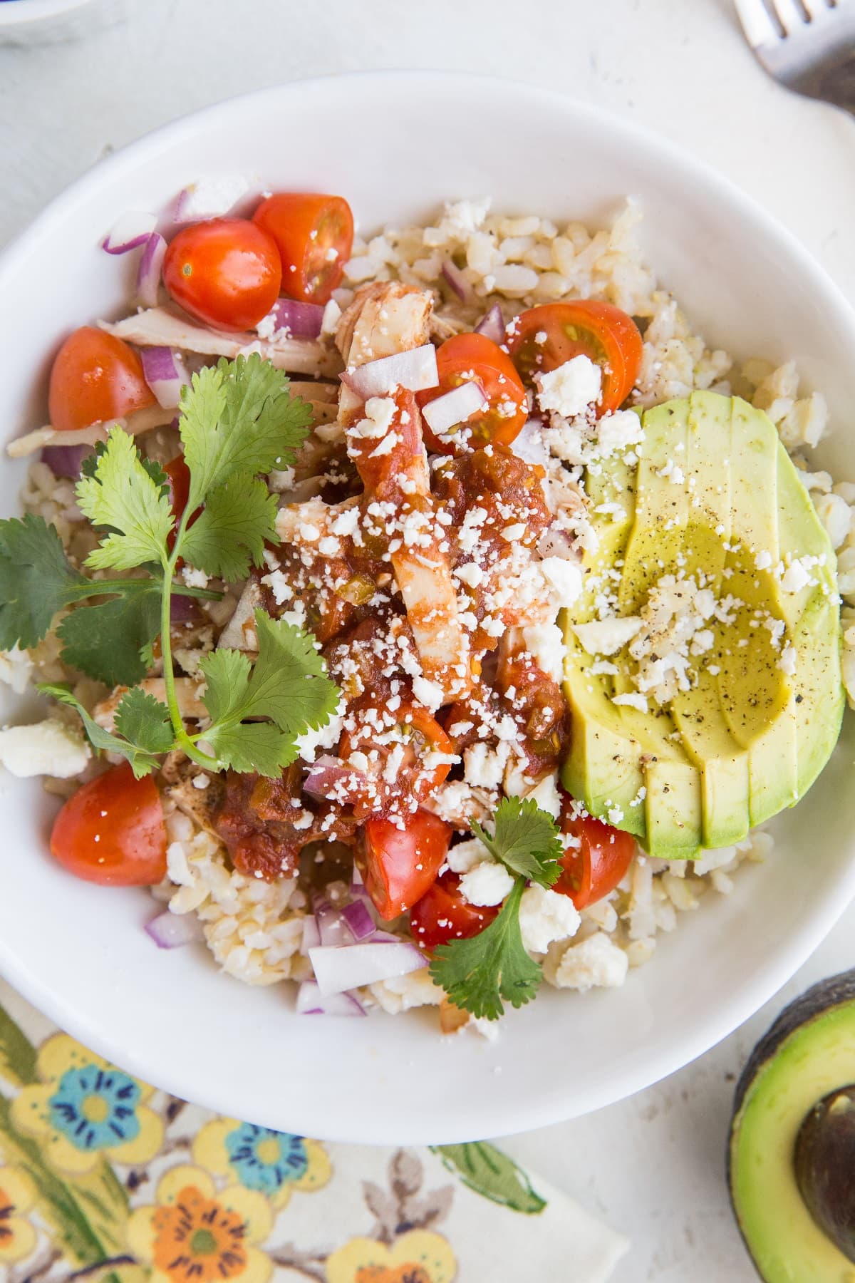 Big Chicken Burrito Bowl with a flower napkin and avocado to the side. Topped with delicious burrito bowl toppings.