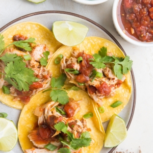 Plate of shredded chicken tacos with cilantro on top and a golden napkin to the side.