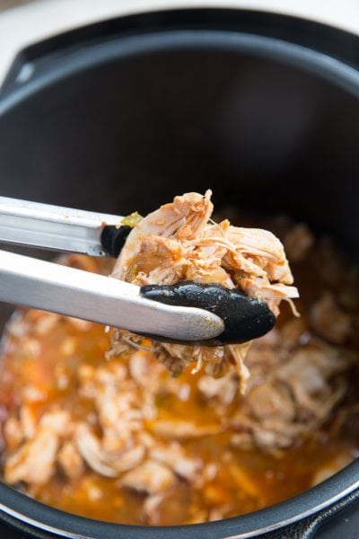 Tongs holding shredded chicken, transferring it back to the pressure cooker with the juices.