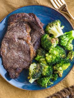 Blue plate with steak and broccoli on it with a gold fork to the side and a golden napkin