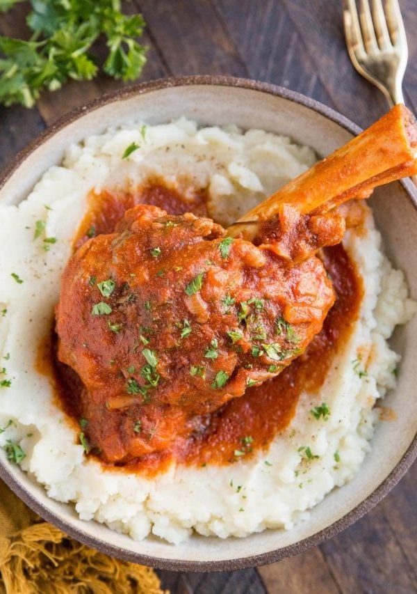 Large bowl with mashed potatoes and lamb shank on top with tomato sauce.