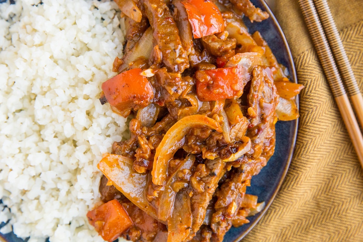 Horizontal image of Beijing beef on a blue plate with cauliflower rice, a brown napkin and chopsticks to the side.