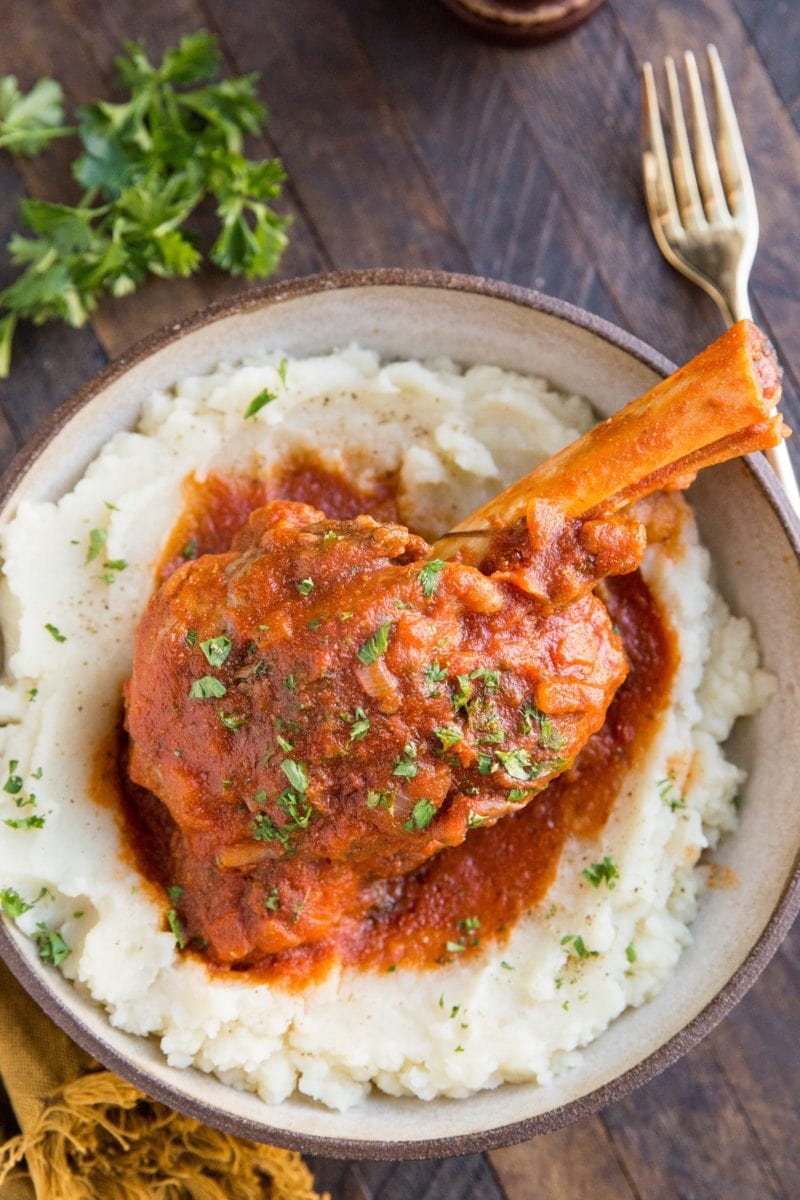 Large bowl with mashed potatoes and lamb shank on top with tomato sauce.
