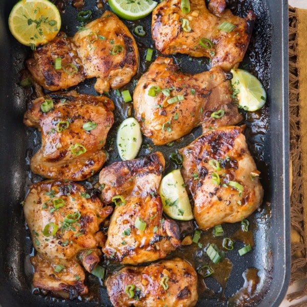 Top down image of whole casserole dish of baked chicken thighs with limes and brown napkin off to the side