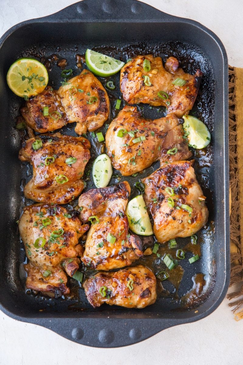 Top down image of whole casserole dish of baked chicken thighs with limes and brown napkin off to the side