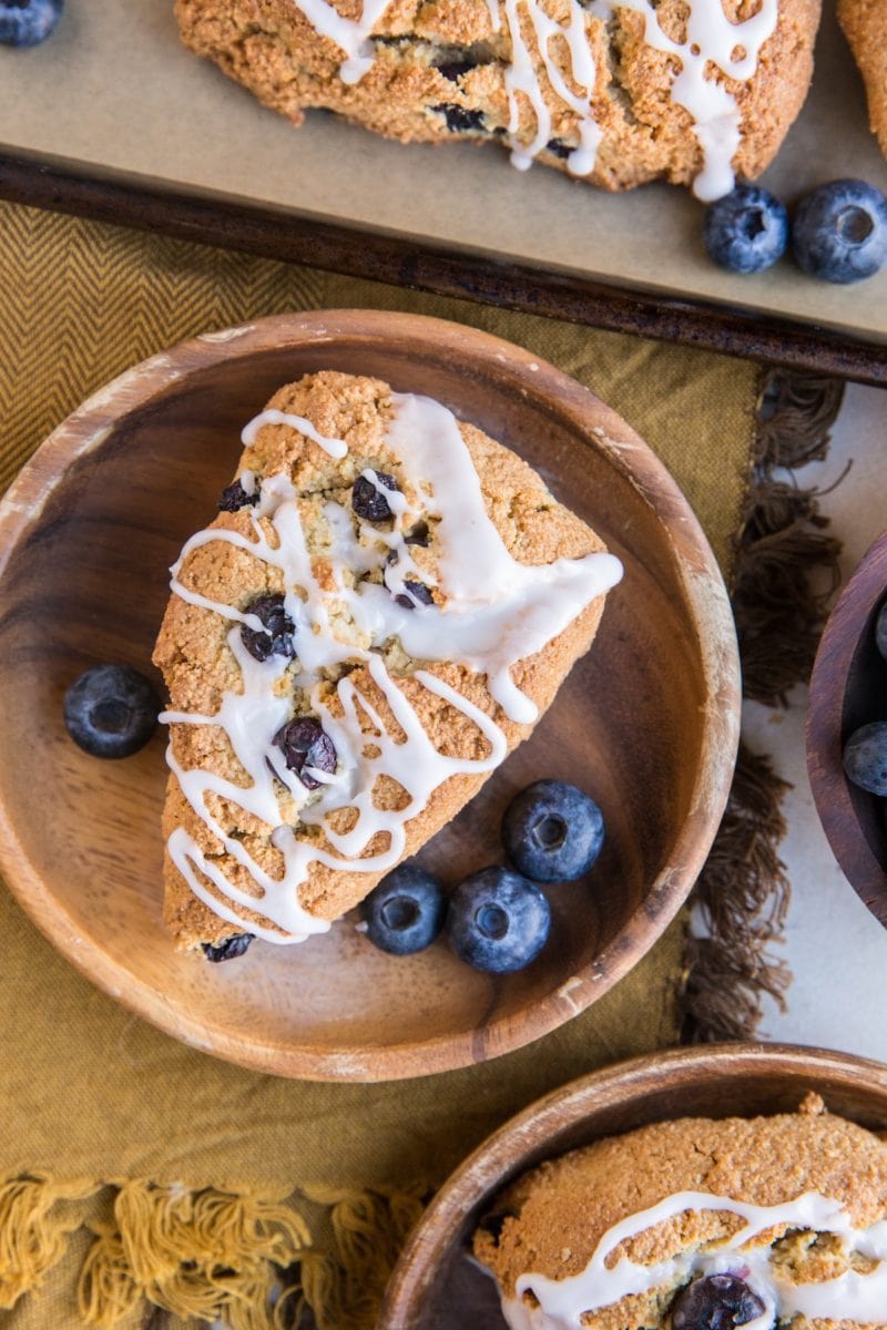 Top down photo of two wooden plates with scones and the baking sheet of the rest of the scones with a brown napkin and fresh blueberries to the sides