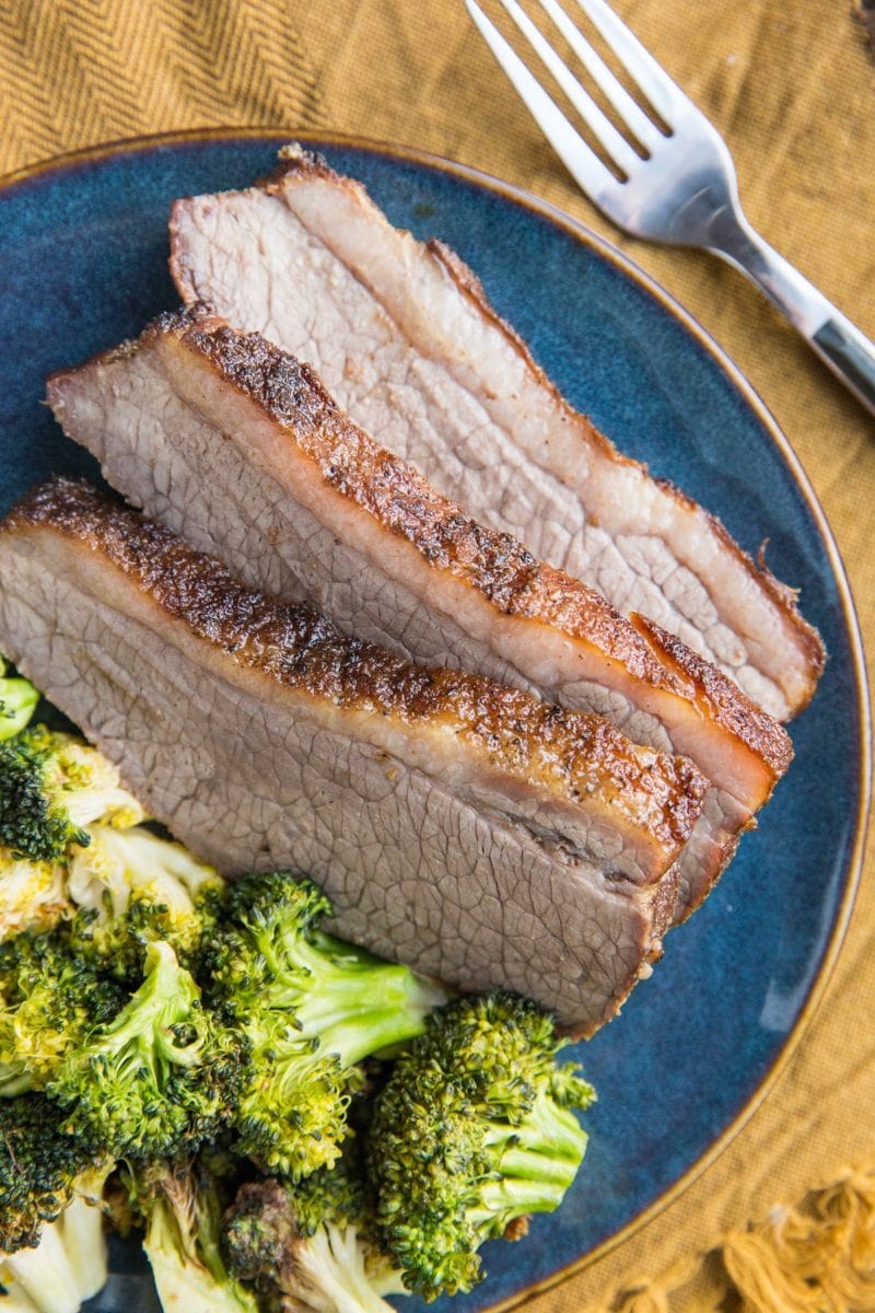 Three thick slices of brisket on a blue plate with roasted broccoli on top of a golden brown napkin