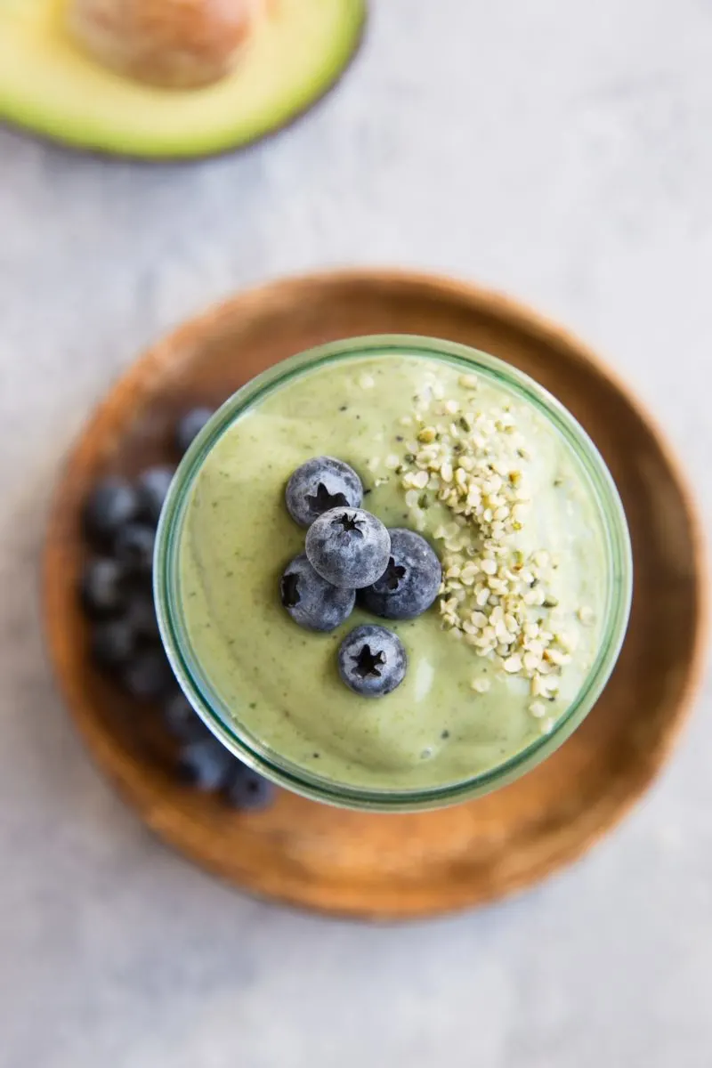 top down photo of avocado smoothie standing on a wooden plate with avocado and blueberries off to the side