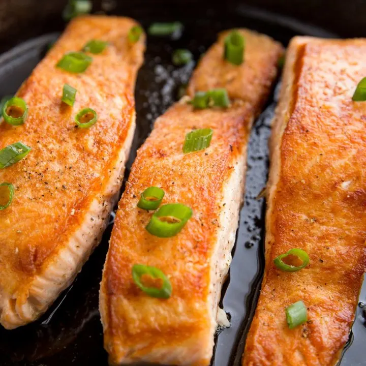 Three salmon filets in a cast iron skillet looking crispy and delicious with green onion sprinkled on top