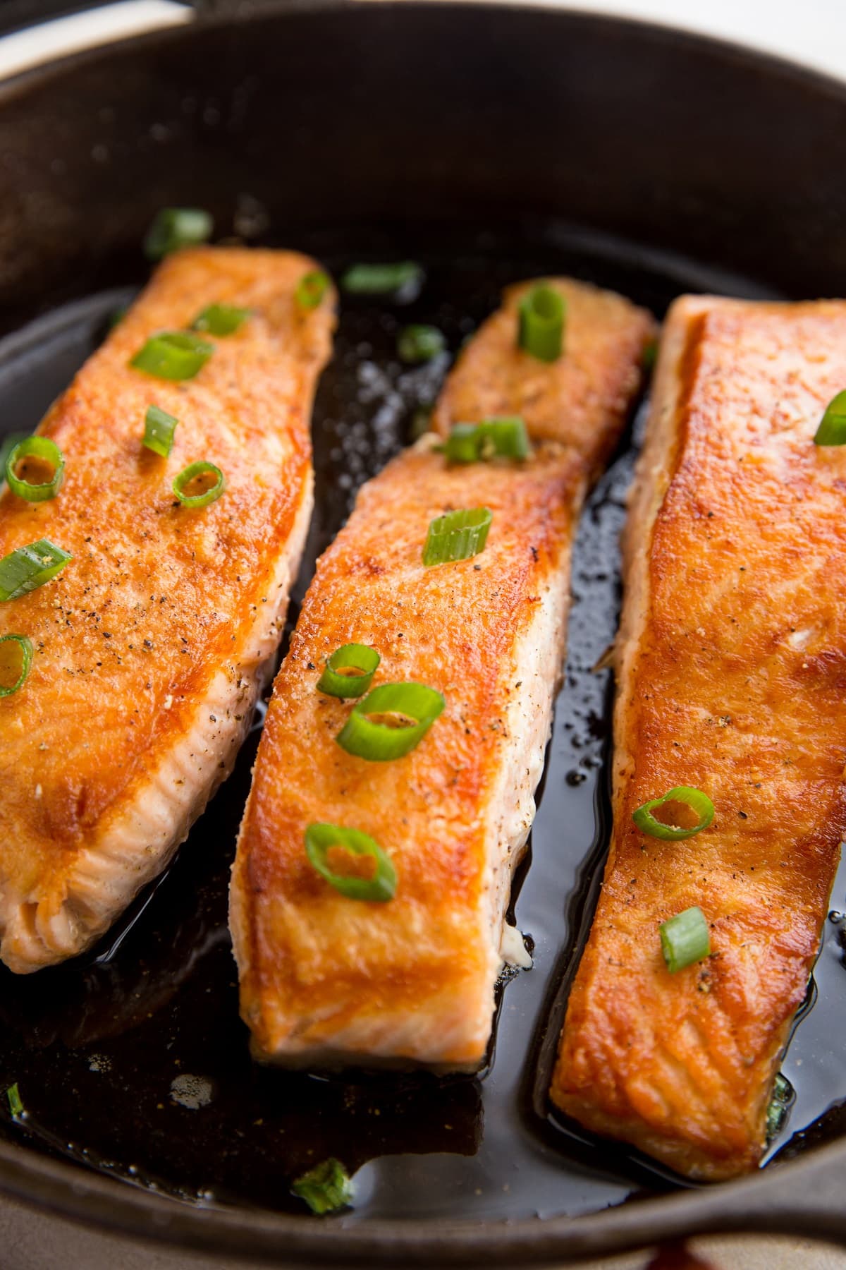 Three salmon filets in a cast iron skillet looking crispy and delicious with green onion sprinkled on top