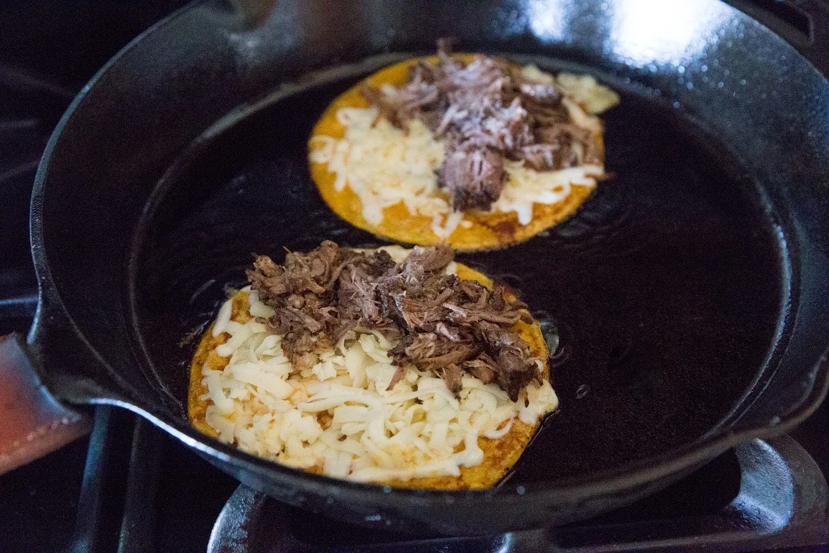 Two corn tortillas in a cast iron skillet with cheese and shredded meat