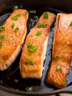 Three salmon filets in a cast iron skillet looking crispy and delicious with green onion sprinkled on top