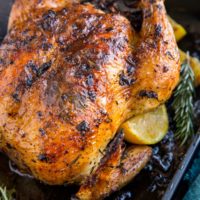 Roasting pan with roast chicken inside and lemon slices and rosemary with a dark blue napkin to the side