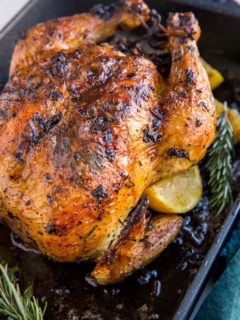 Roasting pan with roast chicken inside and lemon slices and rosemary with a dark blue napkin to the side