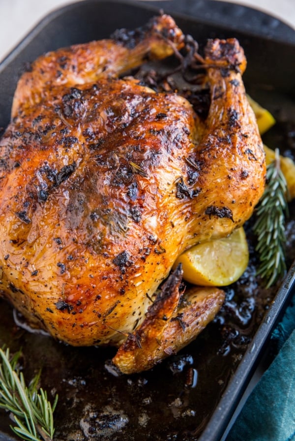 Roasting pan with roast chicken inside and lemon slices and rosemary with a dark blue napkin to the side
