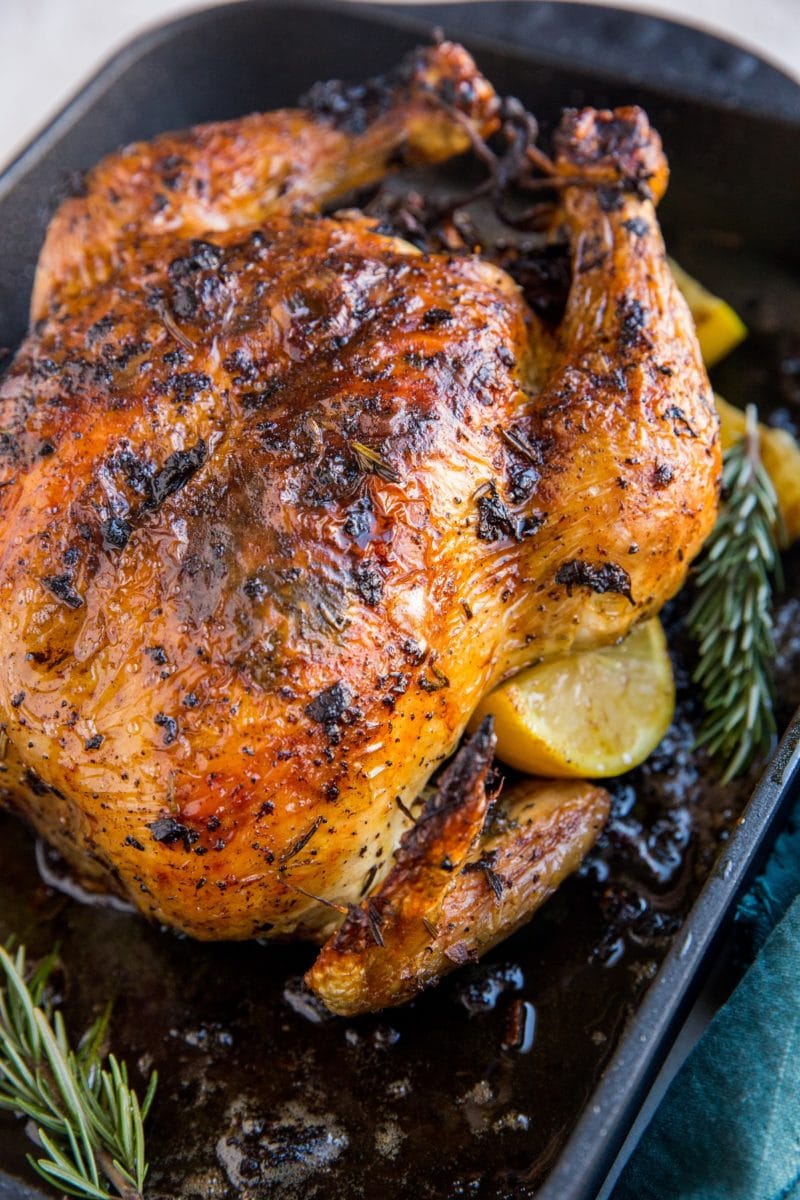 Roasting pan with roast chicken inside and lemon slices and rosemary with a dark blue napkin to the side