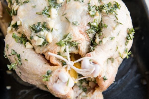Raw whole chicken in a roasting pan with garlic herb butter spread all over it with legs tied with kitchen string and a lemon inside the body cavity.