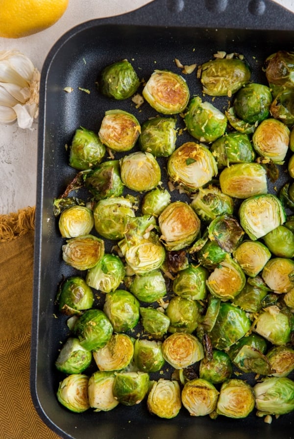 Roasted Brussel Sprouts in a cast iron casserole dish with lemon and garlic off to the side and a brown napkin