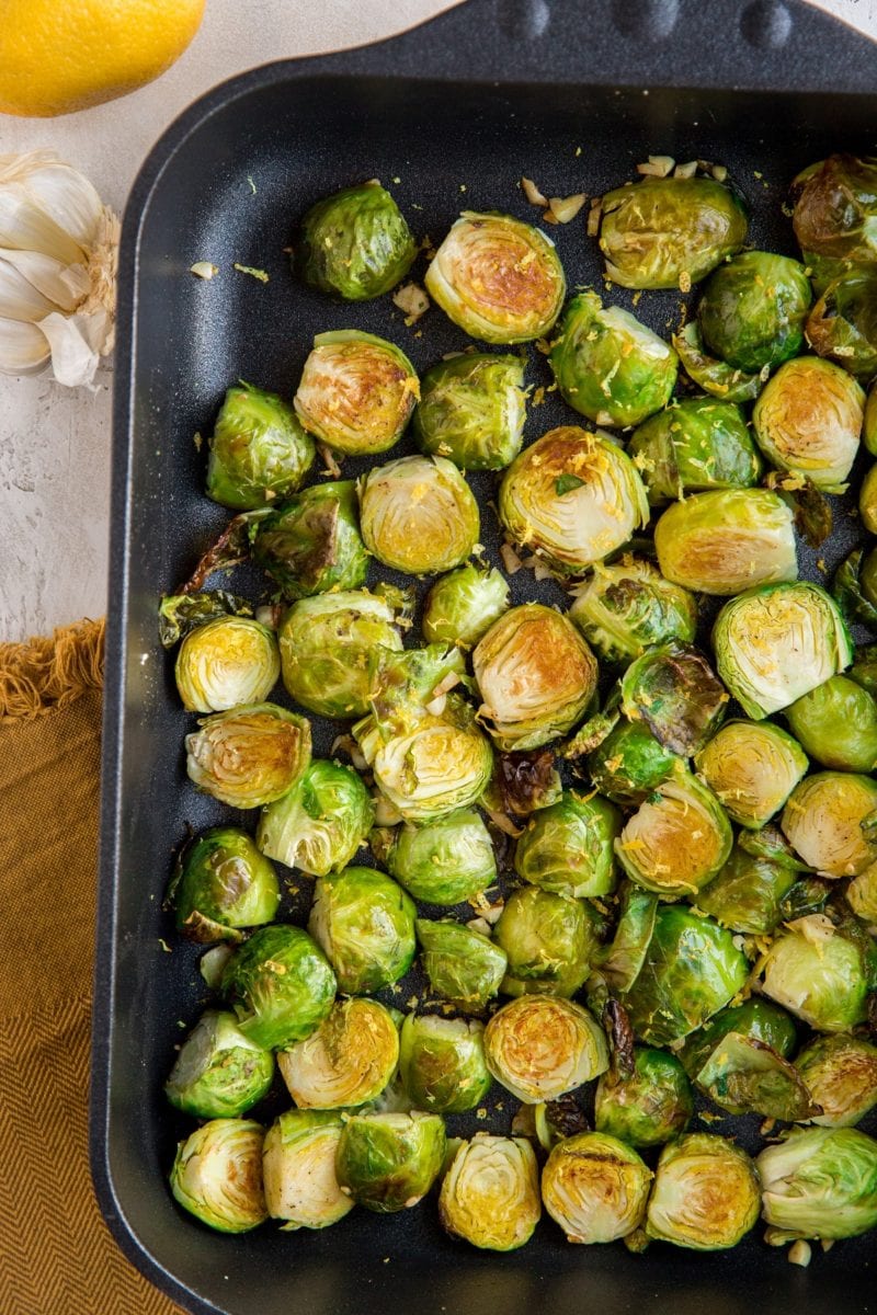 Roasted Brussel Sprouts in a cast iron casserole dish with lemon and garlic off to the side and a brown napkin