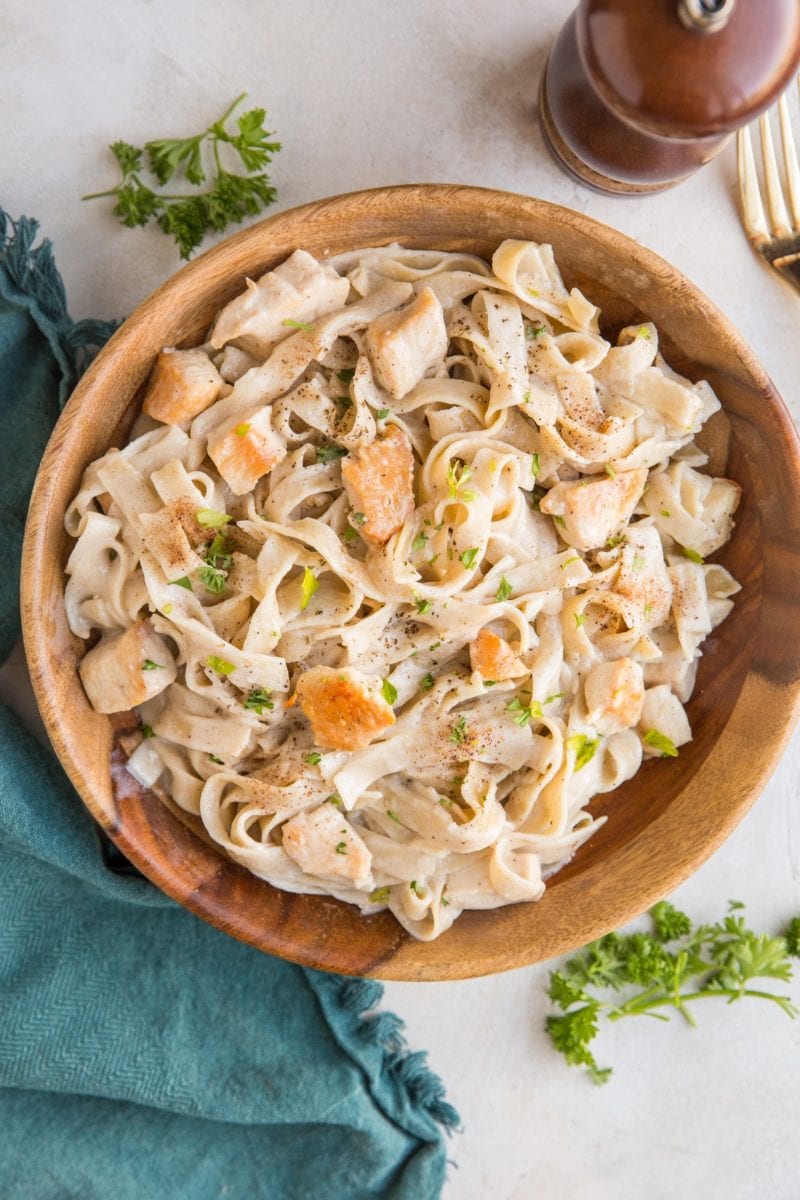 Wooden bowl of gluten-free dairy-free chicken fettuccine alfredo with a side of roasted brussel sprouts and a blue napkin