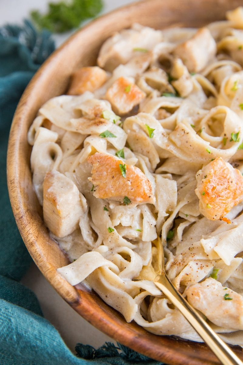 Close up image of creamy chicken fettuccine alfredo in a wooden bowl with a blue napkin and a gold fork