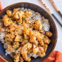 top down photograph of honey garlic chicken over brown rice in a blue bowl with a red napkin and chopsticks off to the side