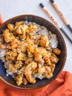 top down photograph of honey garlic chicken over brown rice in a blue bowl with a red napkin and chopsticks off to the side