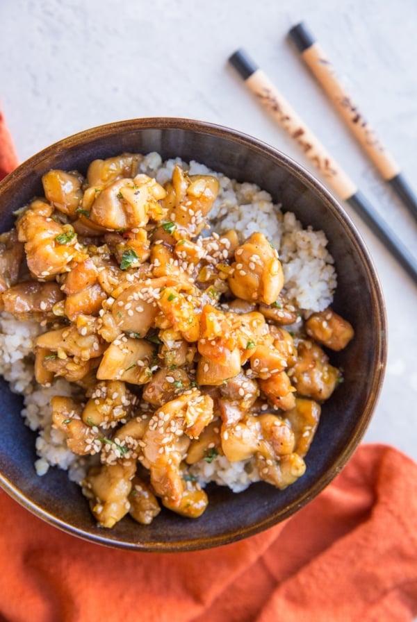 top down photograph of honey garlic chicken over brown rice in a blue bowl with a red napkin and chopsticks off to the side