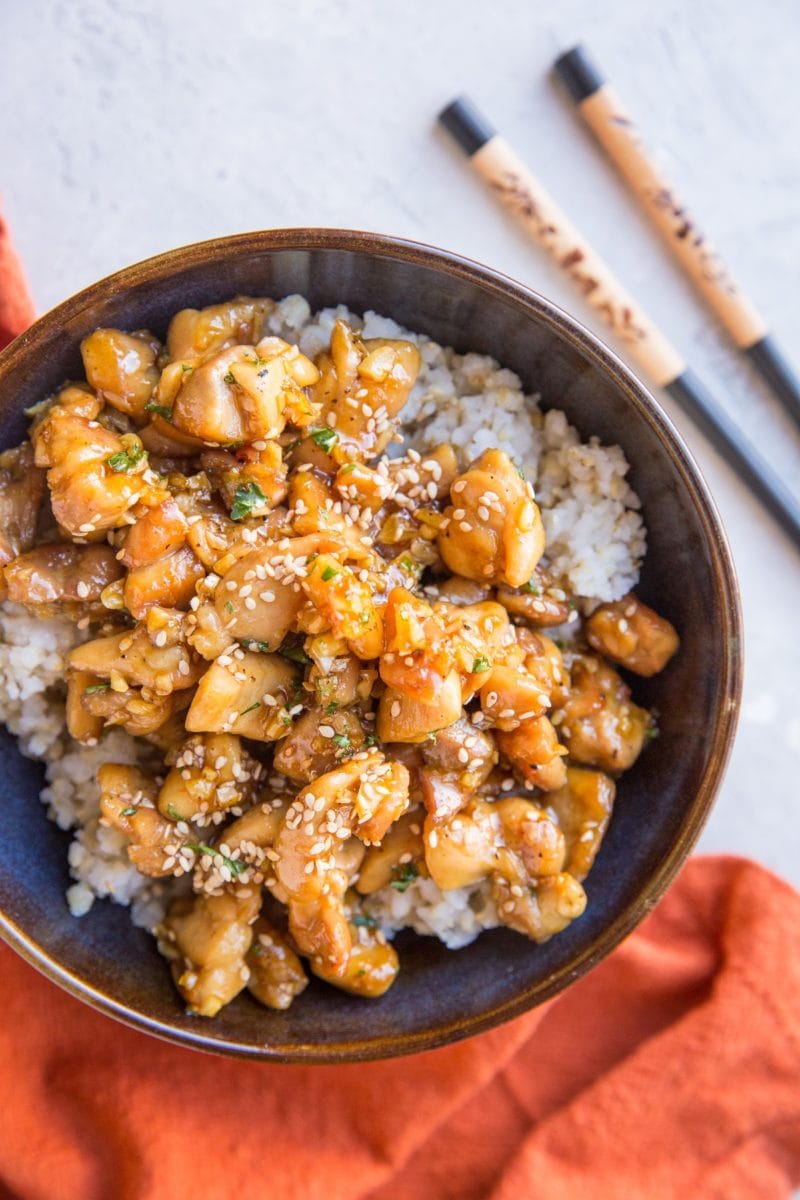 top down photograph of honey garlic chicken over brown rice in a blue bowl with a red napkin and chopsticks off to the side