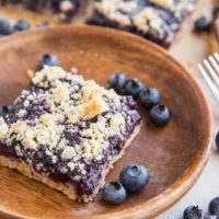 Keto blueberry crumb bars in the background, one bar on a wooden plate in the front with fresh blueberries all around