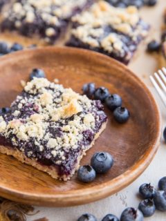 Keto blueberry crumb bars in the background, one bar on a wooden plate in the front with fresh blueberries all around