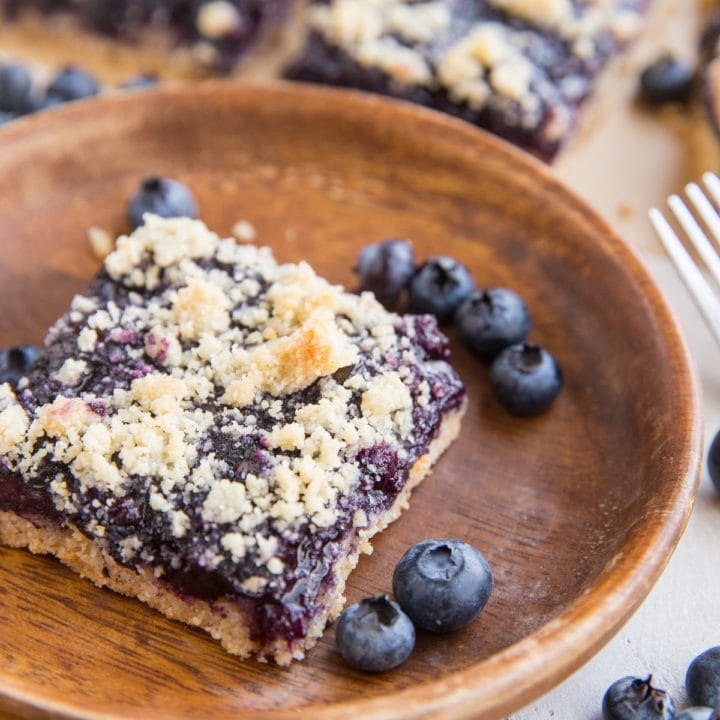 Keto blueberry crumb bars in the background, one bar on a wooden plate in the front with fresh blueberries all around