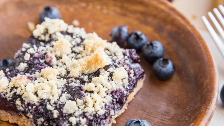 Keto blueberry crumb bars in the background, one bar on a wooden plate in the front with fresh blueberries all around