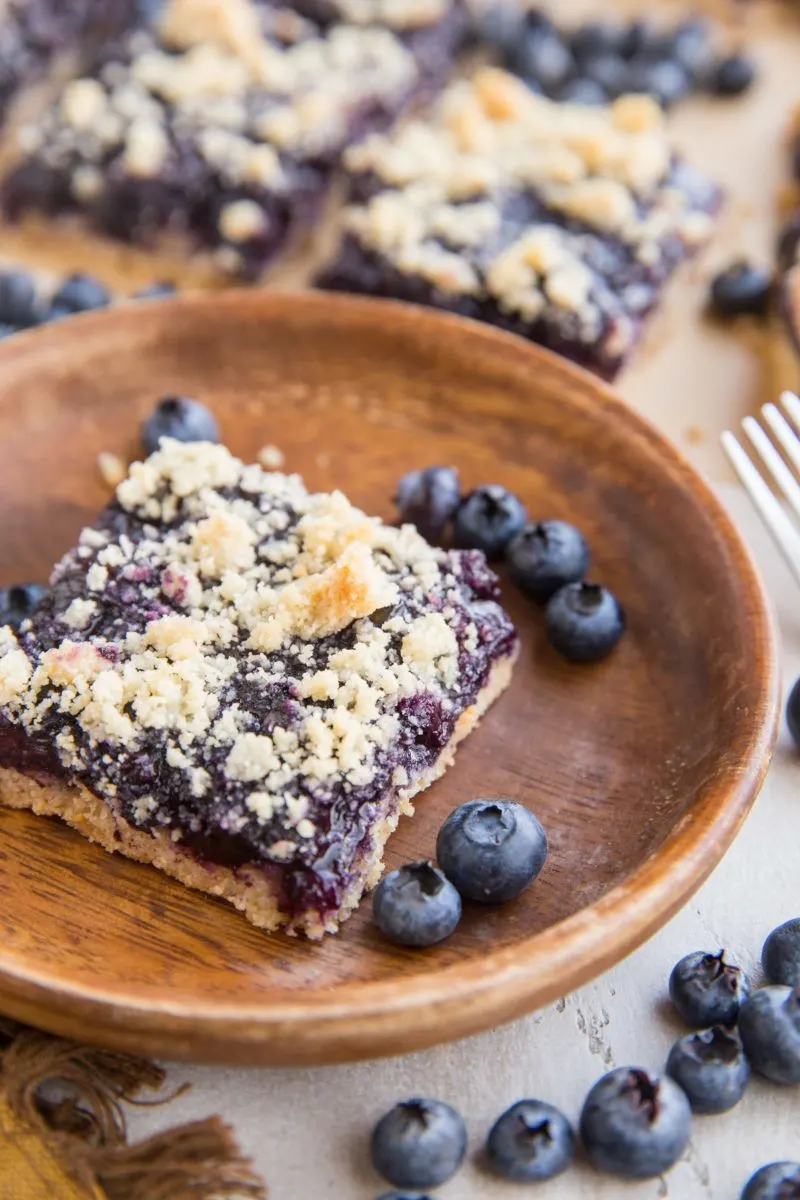 Keto blueberry crumb bars in the background, one bar on a wooden plate in the front with fresh blueberries all around