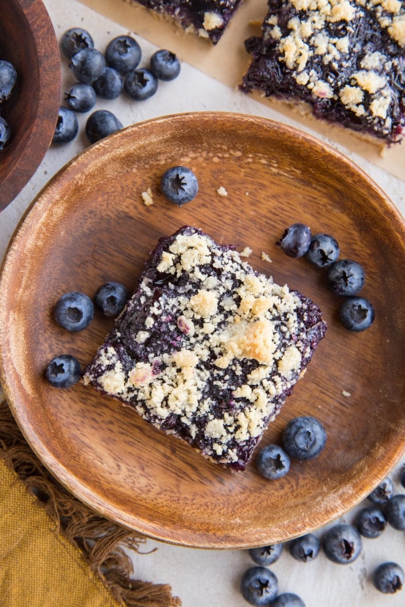 top down photo of a blueberry crumble bar on a wooden plate with fresh blueberries all around