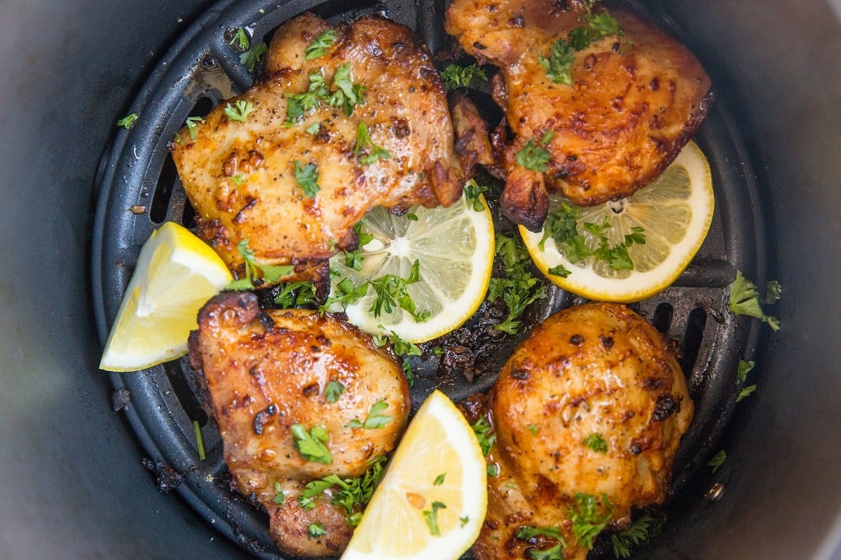 horizontal photo of cooked chicken thighs in an air fryer with slices of lemon and sprinkles of fresh parsley