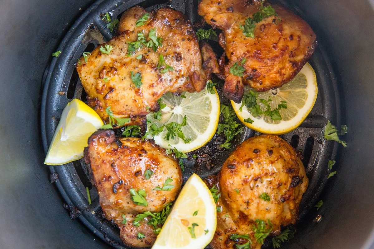 horizontal photo of cooked chicken thighs in an air fryer with slices of lemon and sprinkles of fresh parsley