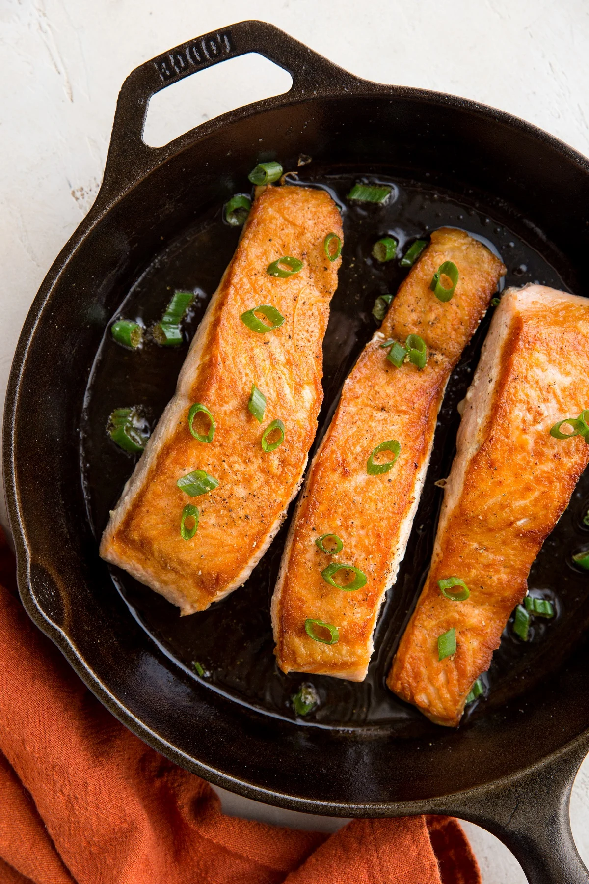 Top down photo of a cast iron skillet with three crispy salmon filets inside, green onion on top and a red napkin off to the side