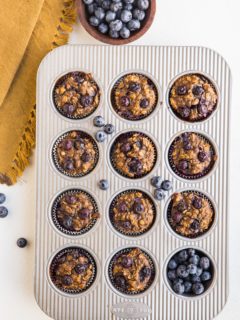 Blueberry Banana Baked Oatmeal Cups in a muffin tray with fresh blueberries to the side and a golden napkin