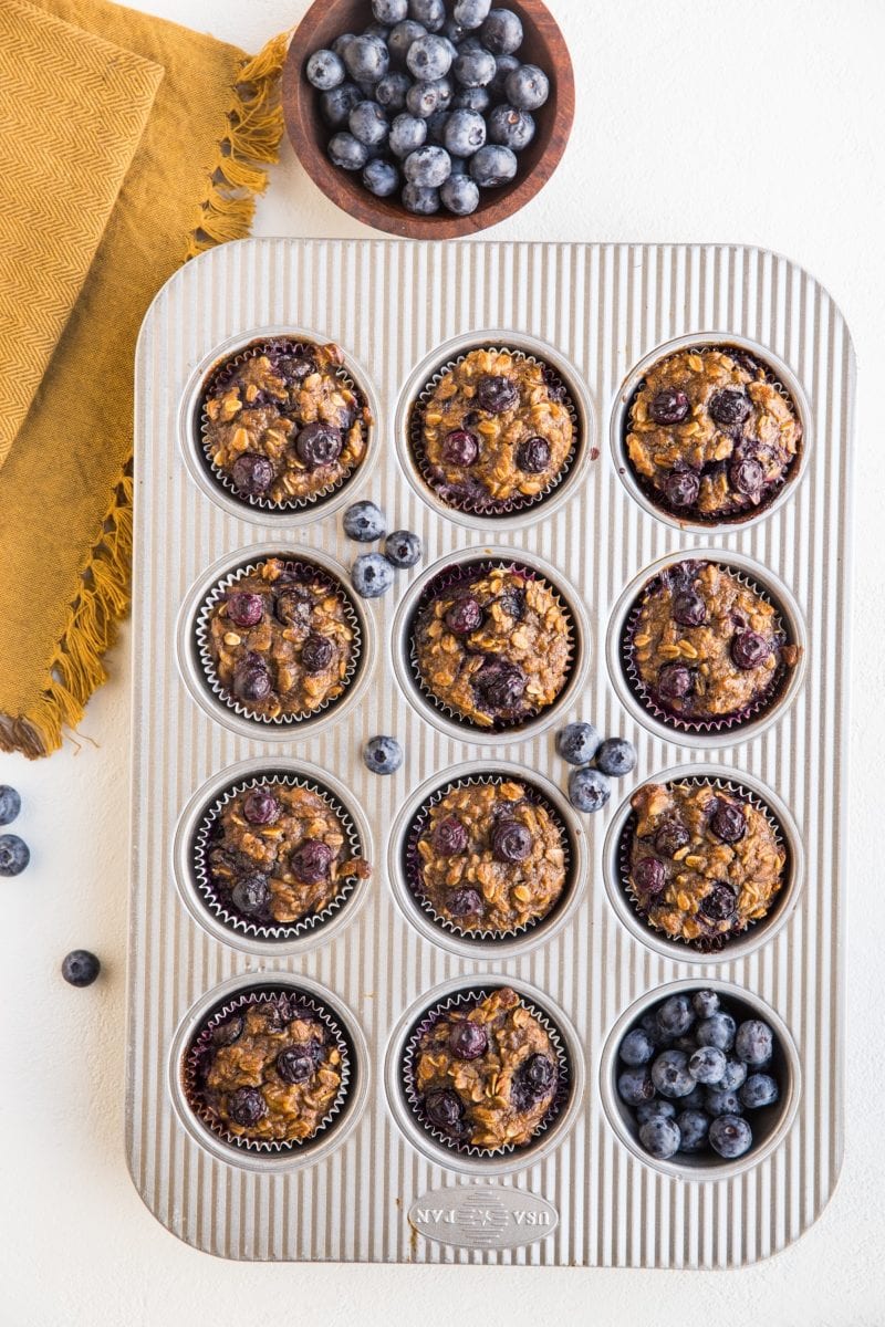 Blueberry Banana Baked Oatmeal Cups in a muffin tray with fresh blueberries to the side and a golden napkin