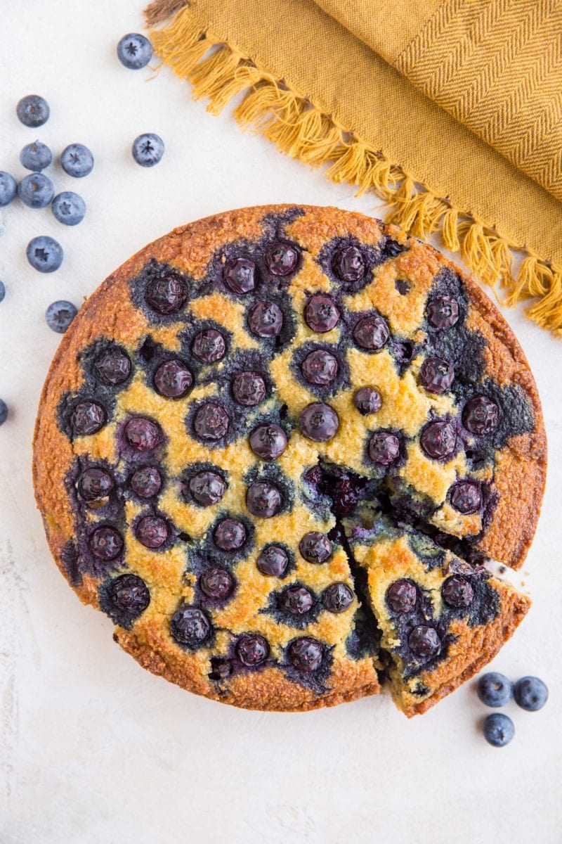 Top down photo of low-carb blueberry cake with one slice cut out, fresh berries to the side and a golden napkin