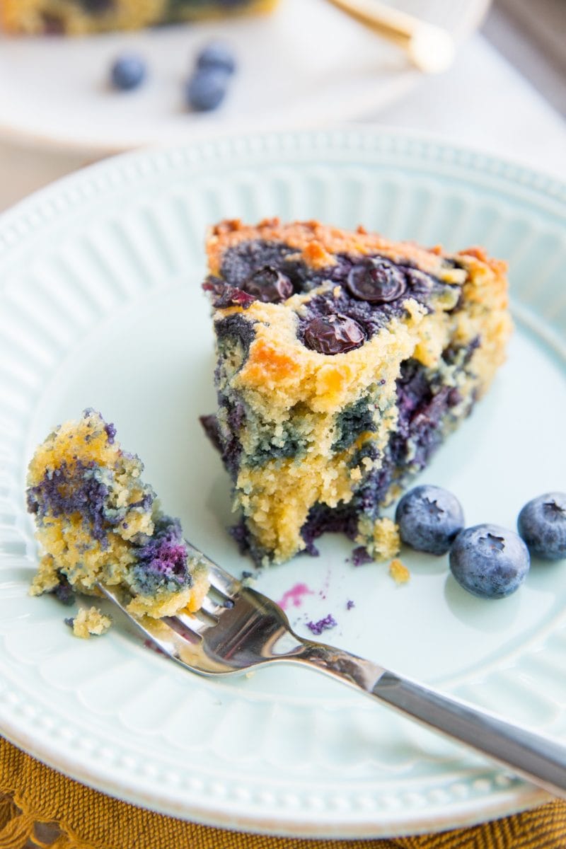 slice of blueberry cake with a bite taken out on a blue plate with fresh blueberries to the side