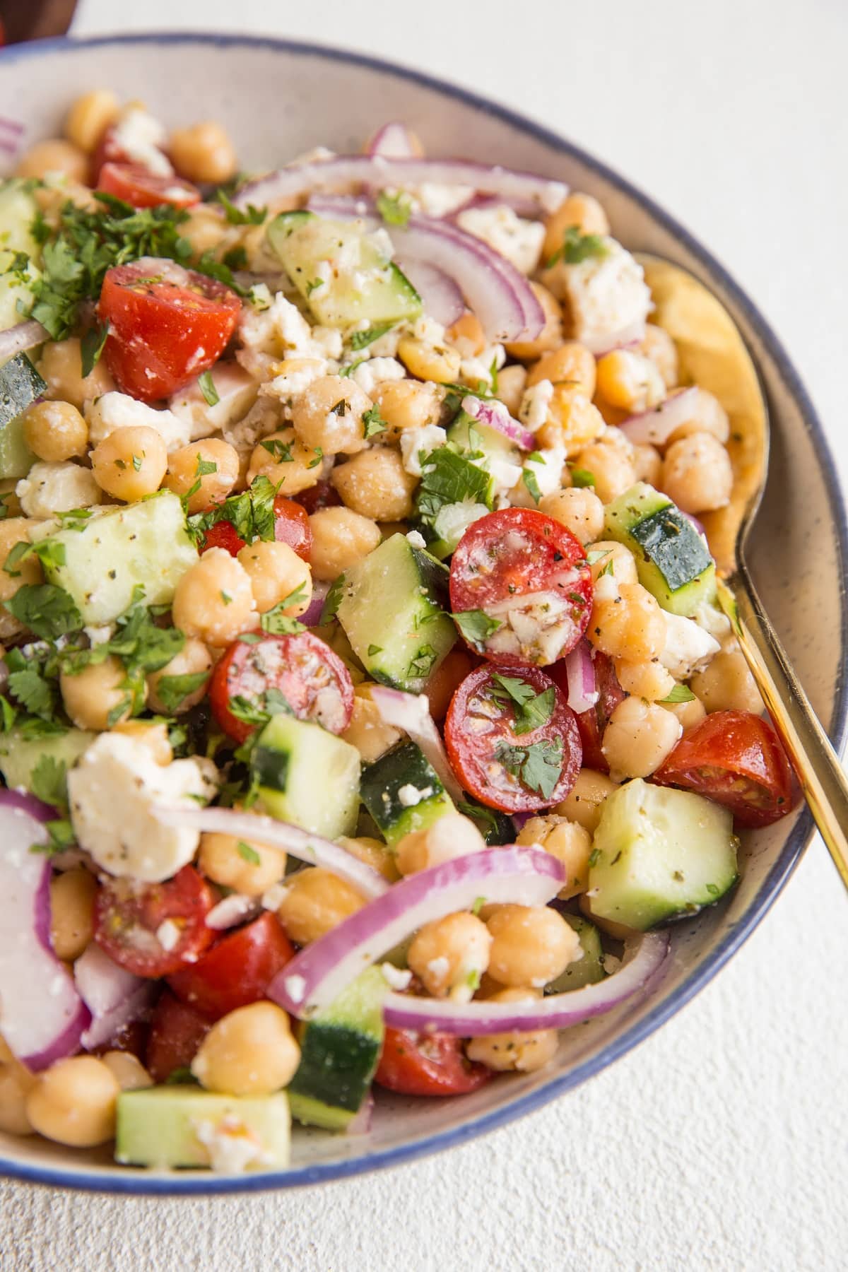 Angled close up shot of garbanzo bean salad in a blue rimmed bowl with a golden spoon.