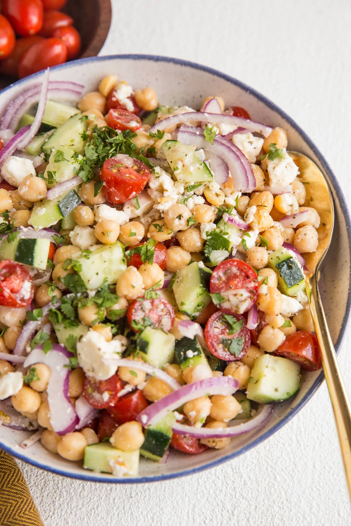 Angle shot of chickpea salad in a bowl with a gold spoon and a wooden bowl of tomatoes