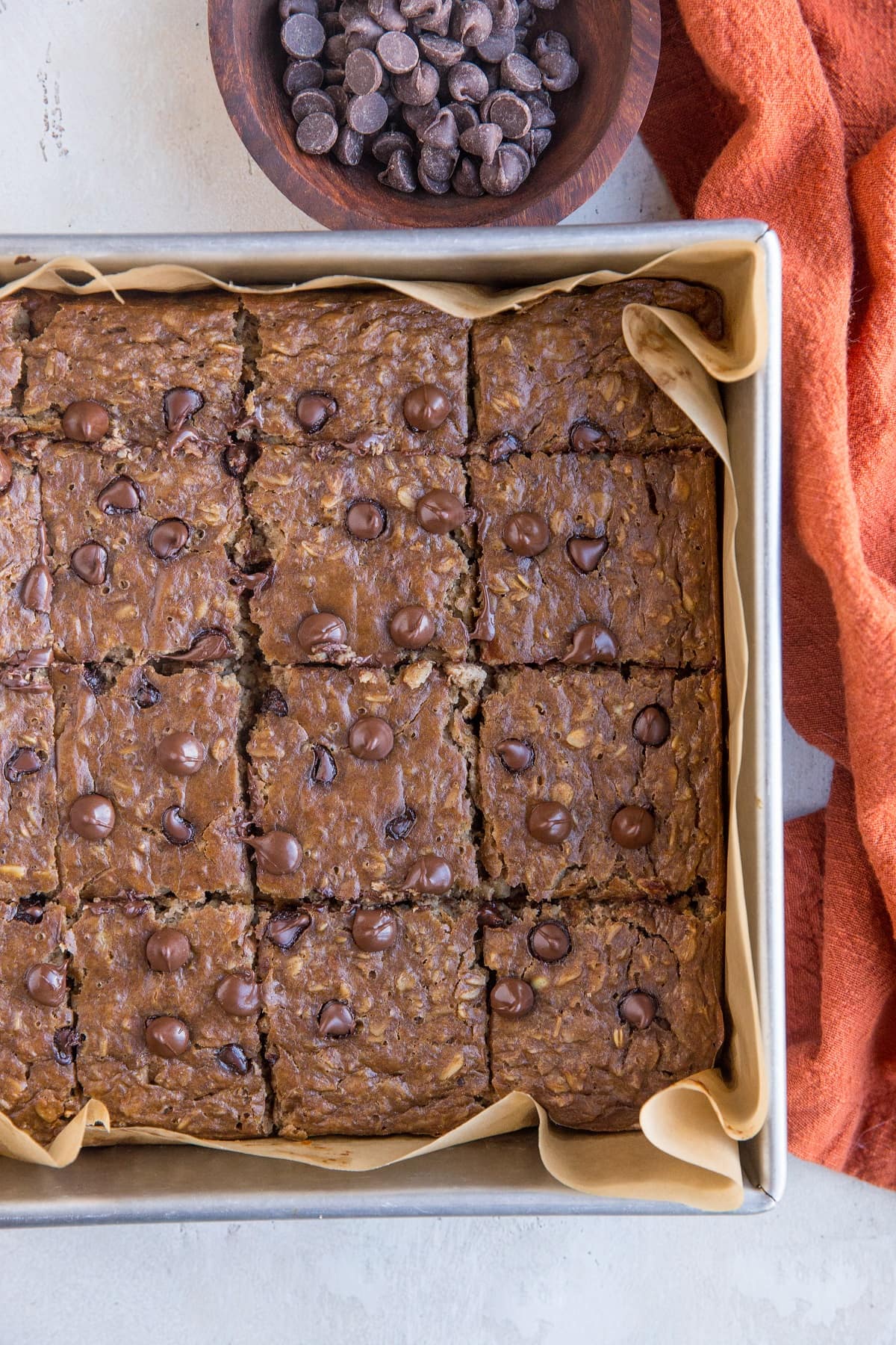 Baking dish of peanut butter banana baked oatmeal with chocolate chips with a red napkin to the side and a bowl of chocolate chips