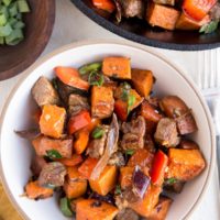 bowl of steak tips with sweet potatoes and bell pepper with a cast iron skillet and a bowl of green onions