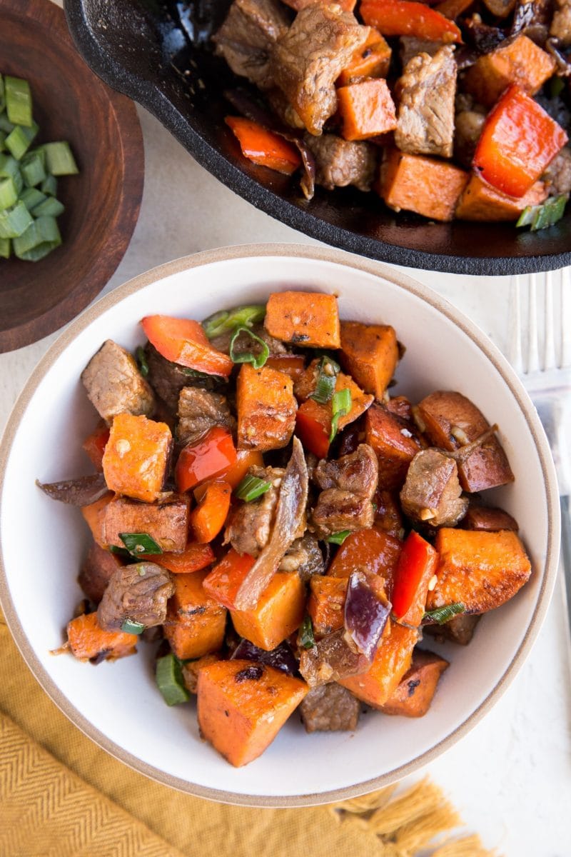 bowl of steak tips with sweet potatoes and bell pepper with a cast iron skillet and a bowl of green onions