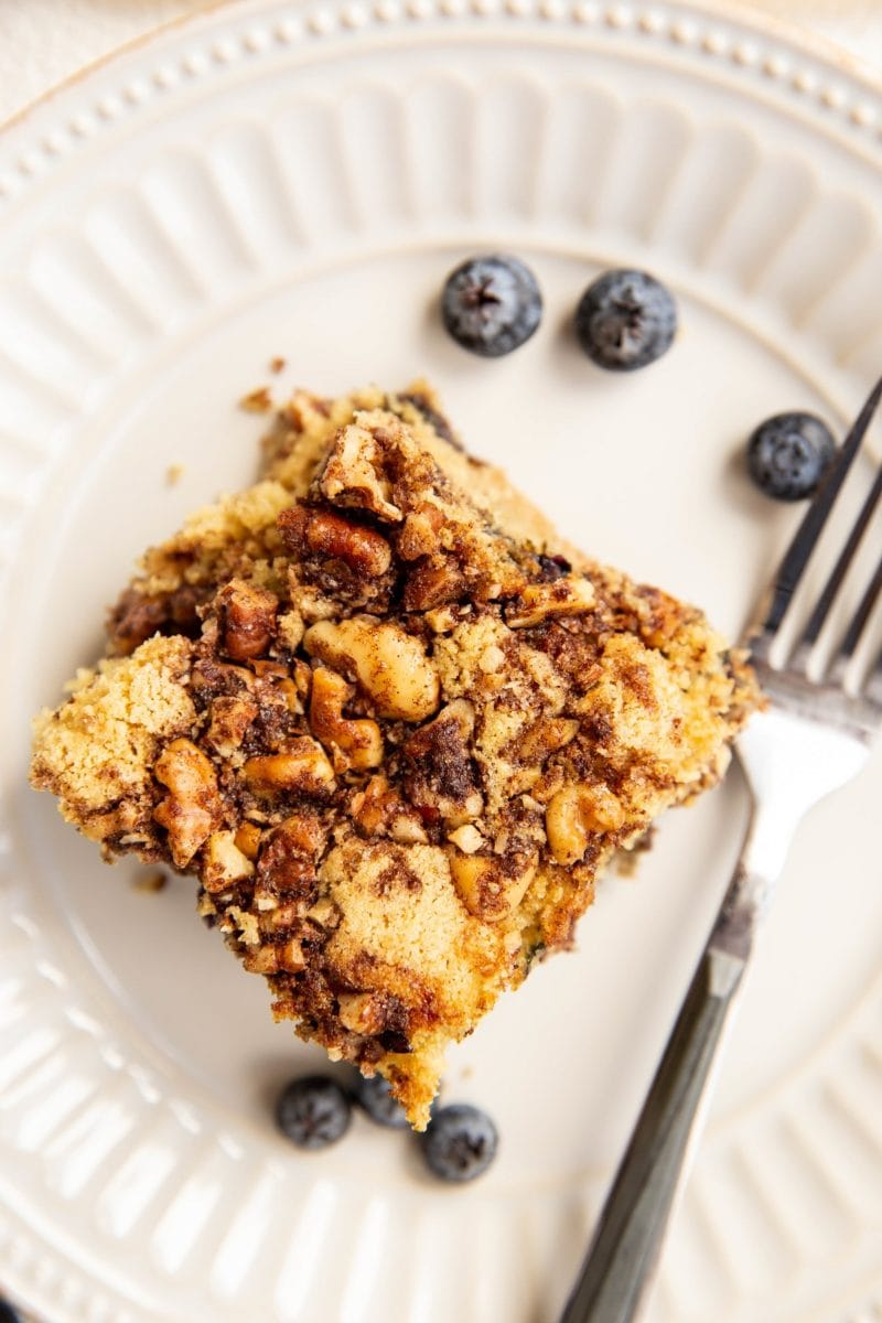 Slice of blueberry coffee cake on a white plate with a fork and fresh blueberries around.